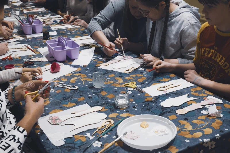 Grupa osób siedzi przy długim stole i wykonuje rękodzieło, malując i wycinając elementy z papieru.