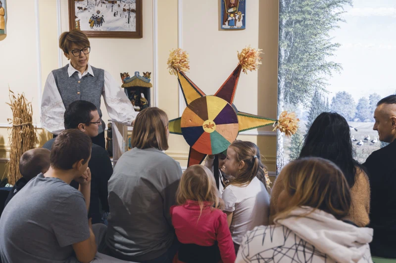 Grupa osób, w tym dzieci, słucha prowadzącej przy kolorowej, gwiaździstej dekoracji.