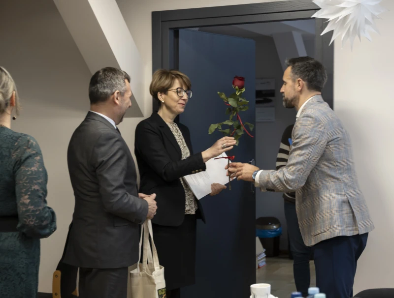 Kobieta odbiera czerwoną różę i dokument od mężczyzny w marynarce, obok stoją inni uczestnicy spotkania.
