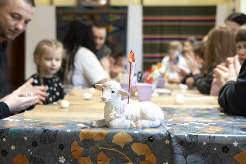 Dzieci i dorośli siedzą przy stole i wspólnie wykonują prace plastyczne z małych elementów.