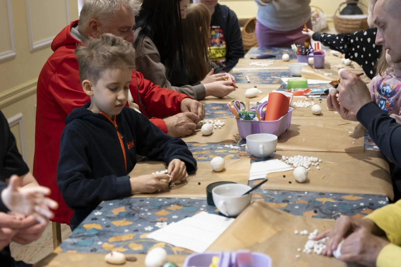 Dzieci i dorośli siedzą przy stole i wspólnie wykonują prace plastyczne z małych elementów.