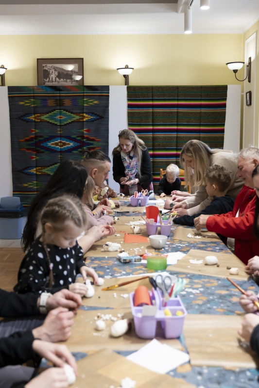 Dzieci i dorośli siedzą przy stole i wspólnie wykonują prace plastyczne z małych elementów.