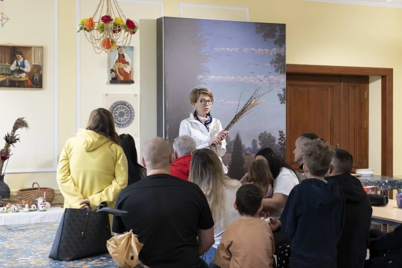 Kobieta stojąca przed grupą dzieci i dorosłych pokazuje gałązki podczas warsztatów rękodzielniczych.