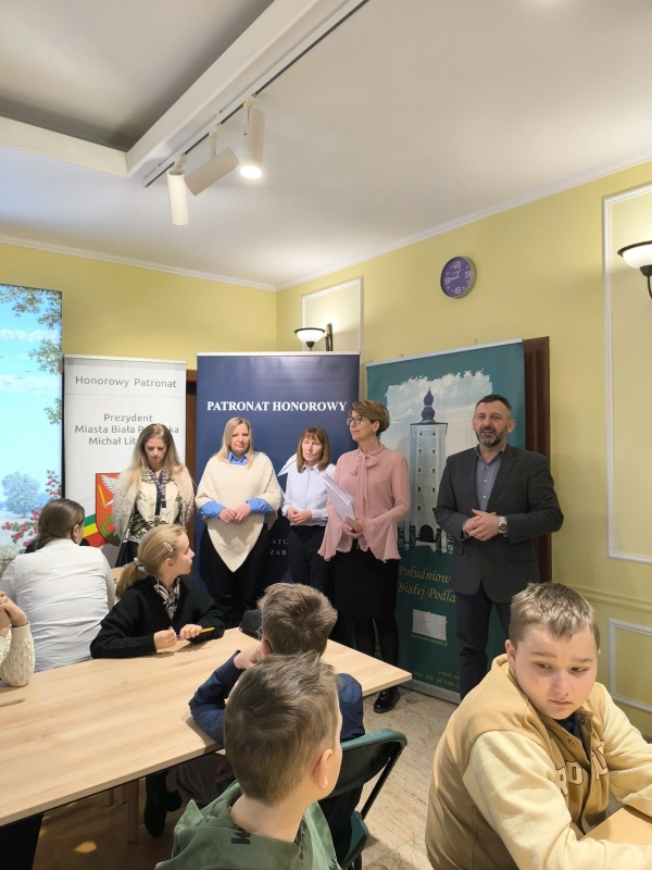 Grupa dorosłych osób stojących przed uczniami w sali ; uczniowie siedzą przy stołach i słuchają.