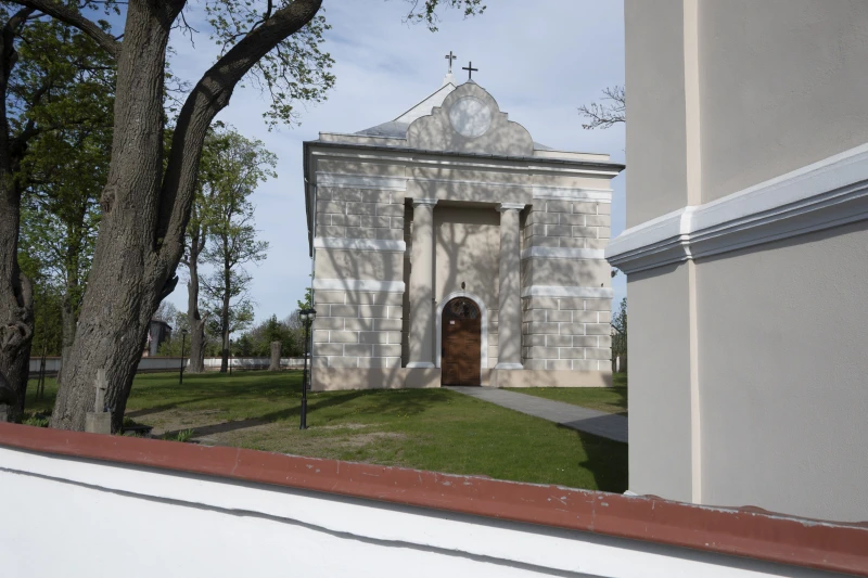 Biała kaplica cmentarna z krzyżem na dachu, otoczona drzewami i nagrobkami, sfotografowana w słoneczny dzień pod błękitnym niebem.