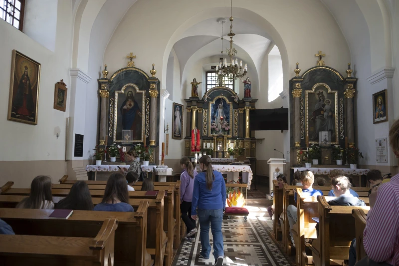 Wnętrze niewielkiego kościoła z ołtarzem i obrazami religijnymi, w którym dzieci siedzą w drewnianych ławkach podczas spotkania.