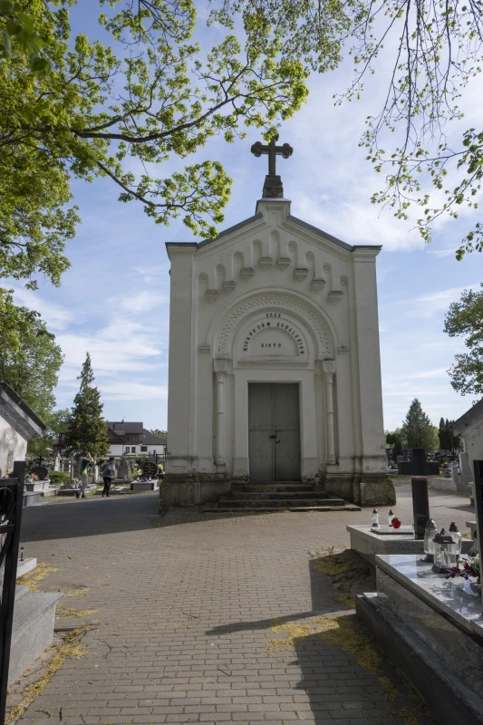 Biała kaplica cmentarna z krzyżem na dachu, otoczona drzewami i nagrobkami, sfotografowana w słoneczny dzień pod błękitnym niebem.