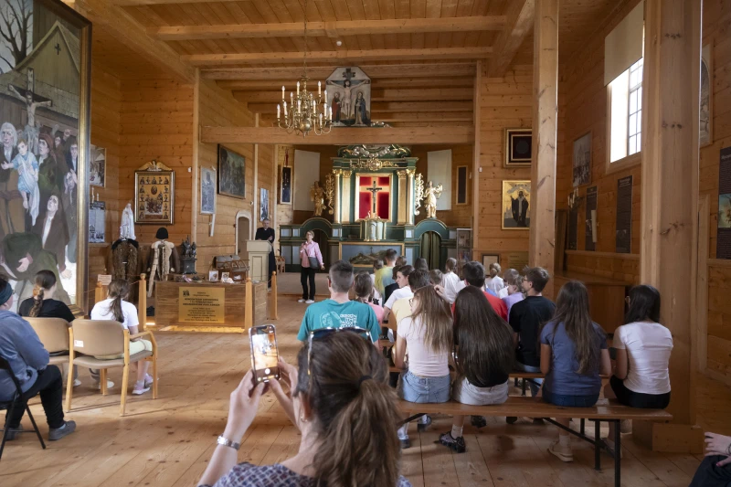 Wnętrze drewnianego kościoła z ołtarzem i religijnymi obrazami. Na ławkach siedzi grupa dzieci i młodzieży uczestniczących w spotkaniu lub lekcji.