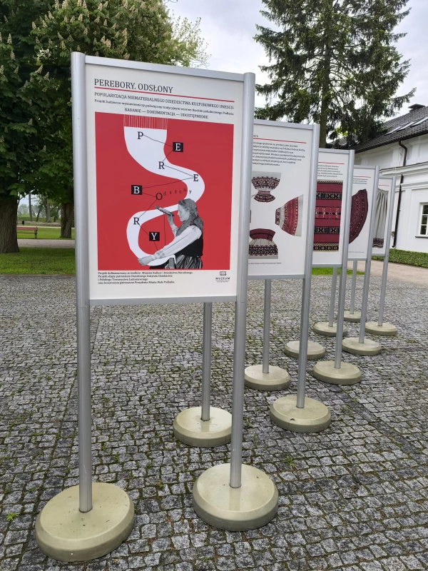 Plansze wystawy plenerowej na brukowanym dziedzińcu, z czerwono-białym plakatem „PEREBORY. ODSŁONY” na pierwszym planie