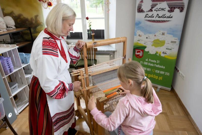 Kobieta w stroju ludowym pokazuje dziecku tkanie na tradycyjnym krosnie.