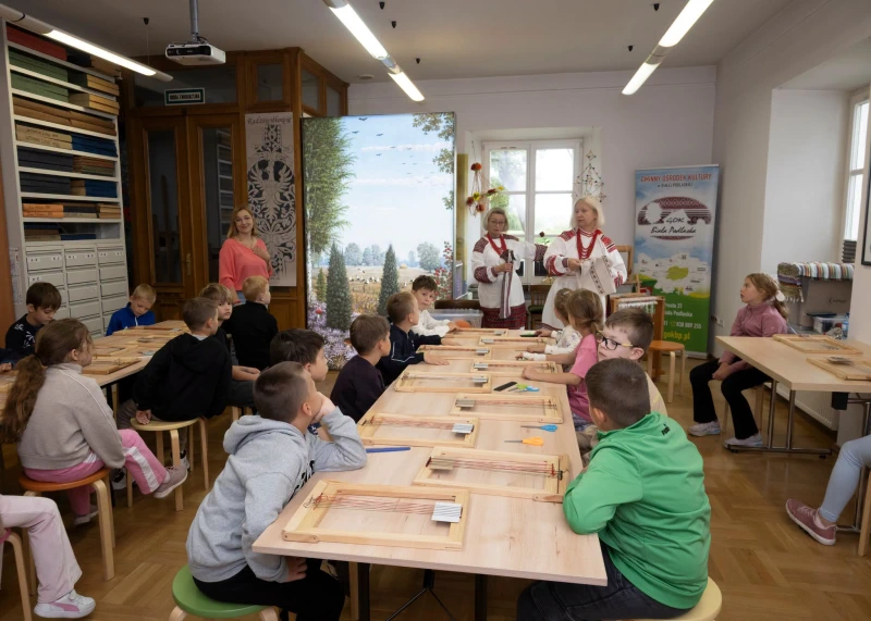 Dziei siedzące przy stołach słuchający wykładu z tkactwa. w tle dwie kobiety w strojach ludowych