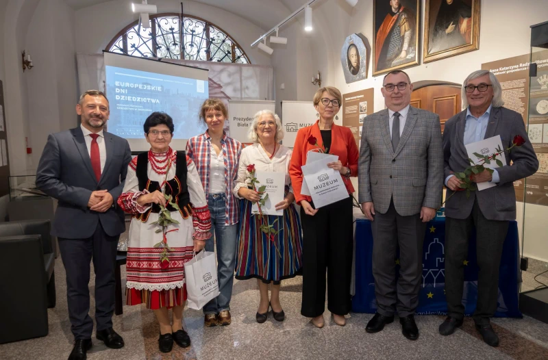 Grupa osób podczas wydarzenia w muzeum, w tym kobieta w stroju ludowym, z kwiatami